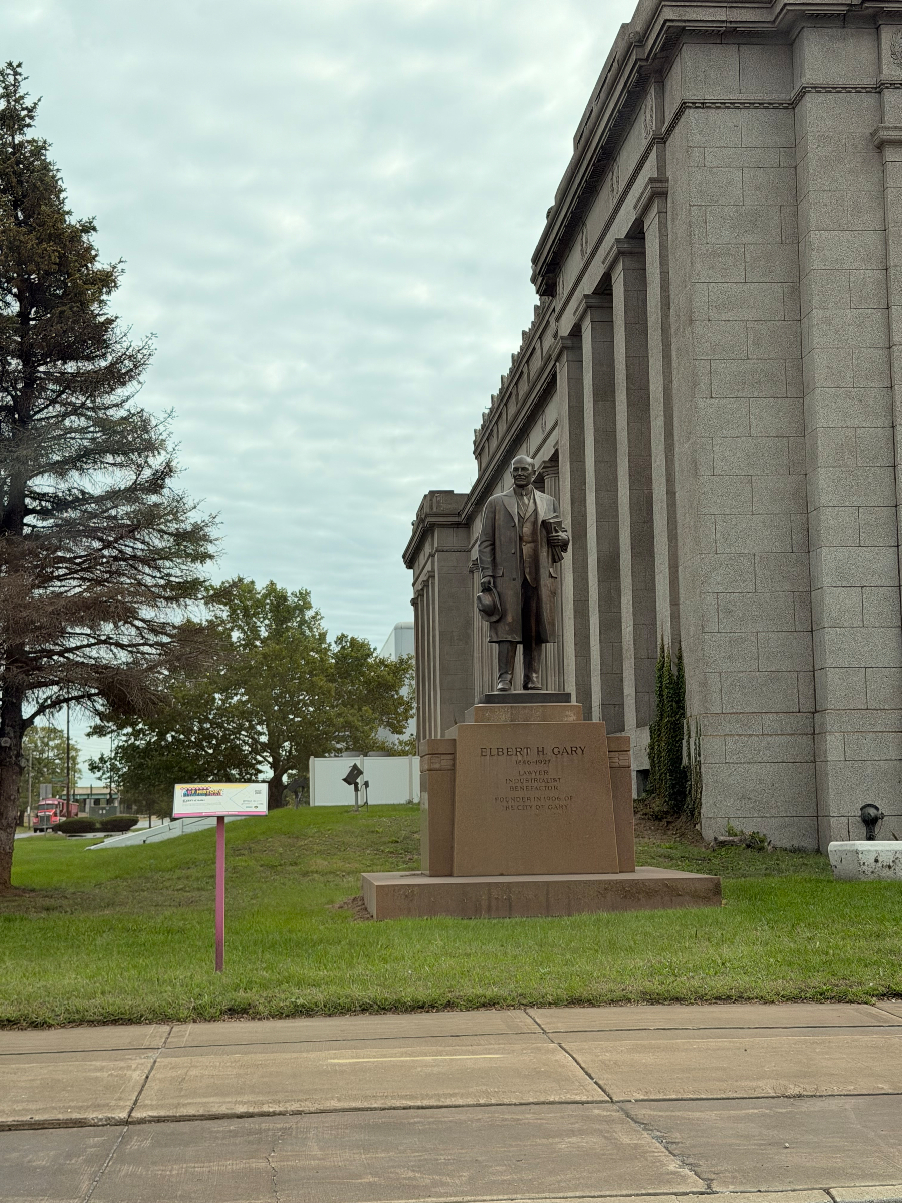 A statue of Elbert H. Gary stands prominently on a pedestal in front of a large stone building with columns, on Broadway Street in Gary, IN, surrounded by grass and trees under a cloudy sky.