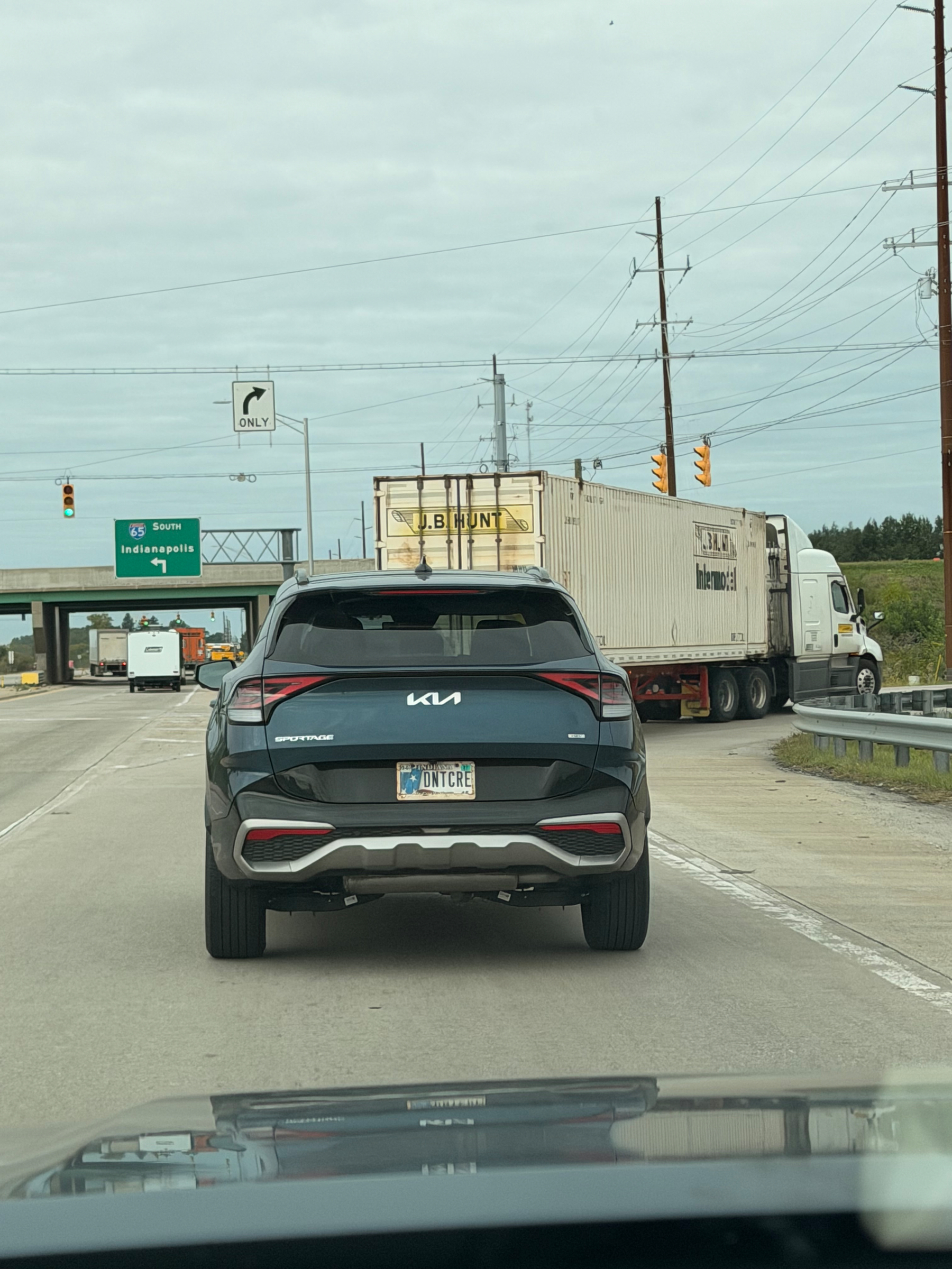 A black Kia SUV with a personalized license plate saying DNT CARE is stopped at a traffic light on a highway near a sign indicating the route to Indianapolis.